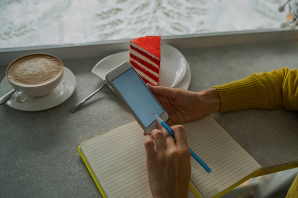 Handy in den Händen einer Frau, die SMS schreibt, im Café sitzt. geschnittenes Bild von Händen an einem Tisch mit Kaffee und Kuchen. Draufsicht einer Frau, die ihr Smartphone in einem Café benutzt. Getöntes Image. - Foto, Bild