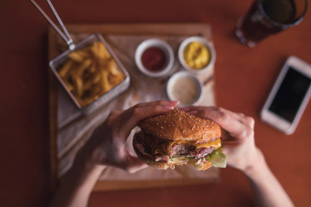 Hands holding fresh delicious burgers with french fries, sauce and drink on the wooden table top view. Delicious hamburger on wooden board. Hamburger with lettuce and cheese. Fast food. Toned image. - Фото, изображение