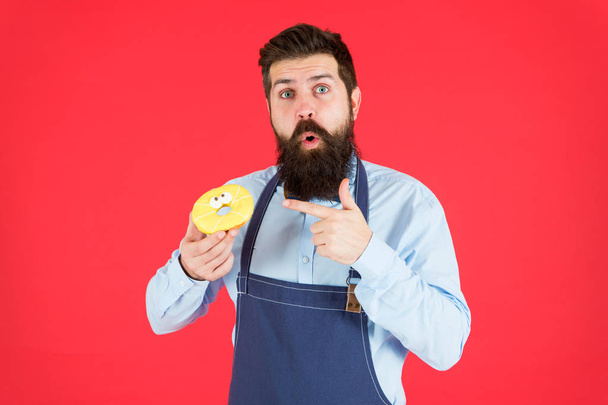 Sweet donut from baker. Man bearded baker in cooking apron hold cute dessert. Ways to reduce hunger and appetite. Hipster bearded baker hold glazed donut on red background. Cafe and bakery concept - Foto, Imagen
