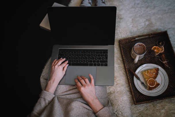 Top view of young woman sitting on floor with plate cakes and laptop. Winter holidays. Online shopping. Christmas, comfort, leisure and people concept. Seasonal sale. Toned image. Selecnive focus. - 写真・画像
