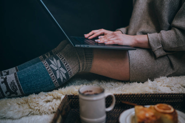 木製の床に座っている少女。ノートパソコンとコーヒーカップウールの靴下の足。女性は居心地の良い家でくつろぐ。冬とクリスマスの休日のコンセプト。選択的な焦点。トーン画像. - 写真・画像