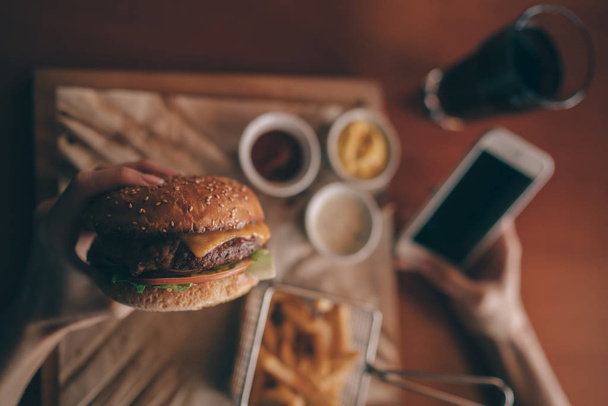 Women's hand using an application to send an sms message in her smartphone device while eating. People and eating concept. Hamburger, french fries, ketchup, mustard on a wooden board. Toned image. - Fotoğraf, Görsel