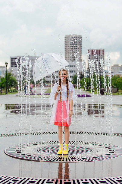 Νεαρό υγρό όμορφο κορίτσι με δύο πλεξούδες σε κίτρινες μπότες και με διάφανη ομπρέλα στέκεται μέσα στο σιντριβάνι. - Φωτογραφία, εικόνα