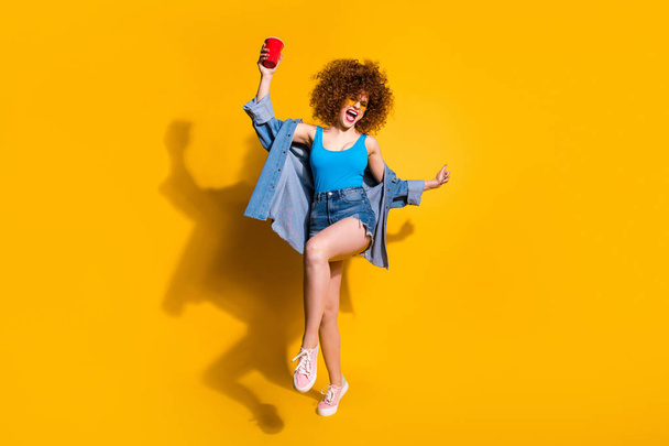 Full length body size photo funny funky she her lady wavy styling curls sing song beverage little drunk hang out wear specs casual jeans denim shirt shorts tank top clothes isolated yellow background - 写真・画像