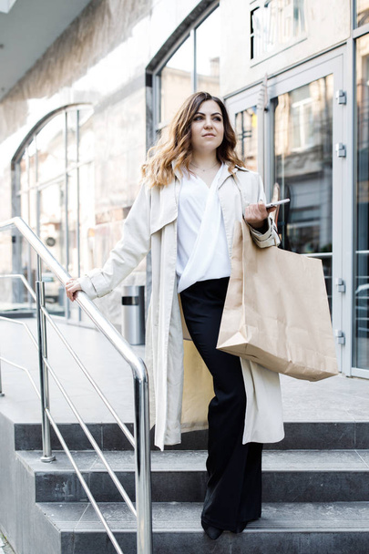 A young, sympathetic woman, not a thin-headed body building, walks around the city with a shopping bag, holds the phone in her hands and has fun. - Foto, Imagen