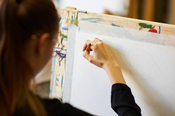 Young beautiful fair-haired girl in glasses dressed in black blouse sits at the easel and draws a picture with a pencil in the drawing school - Fotografie, Obrázek