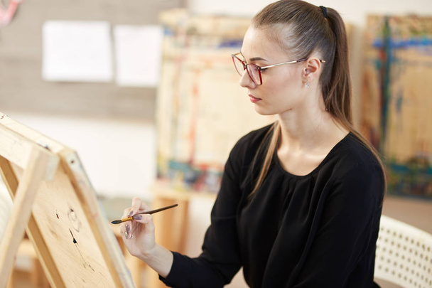 Pretty fair-haired girl in glasses dressed in black blouse sits at the easel and paints a picture in the art studio - Fotografie, Obrázek