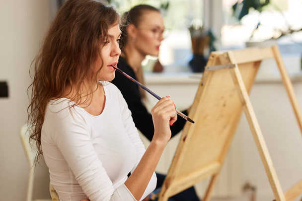 Charming girl with brown curly hair dressed in white blouse creates a picture at the easel holding the brush in her mouth in the drawing school - Photo, Image
