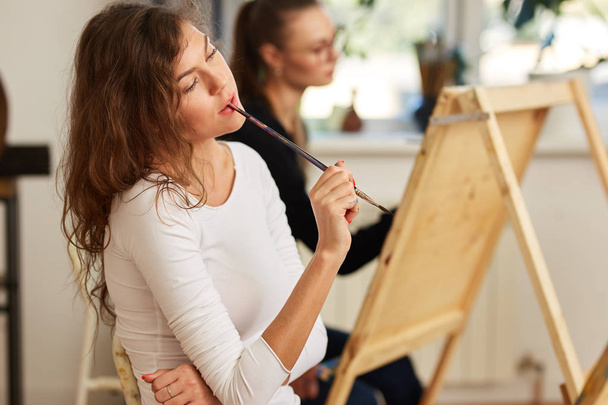 Charming girl with brown curly hair dressed in white blouse creates a picture at the easel holding the brush in her mouth in the drawing school - Foto, Imagem
