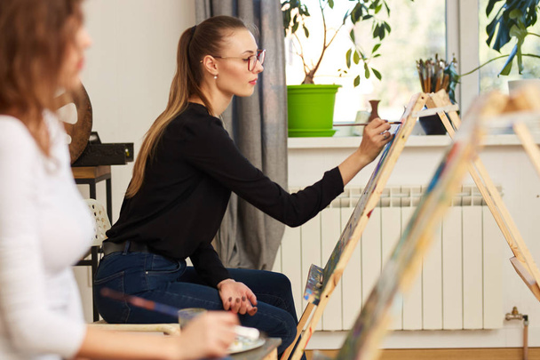 Young beautiful girl in glasses dressed in black blouse and jeans sits at the easel and paints a picture in the drawing school - Foto, Imagem