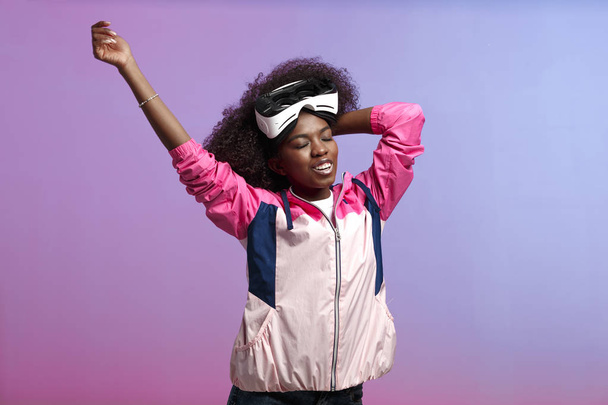 Funny curly brown-haired girl dressed in the pink sports jacket is wearing on her head the virtual reality glasses in the studio on neon background - 写真・画像