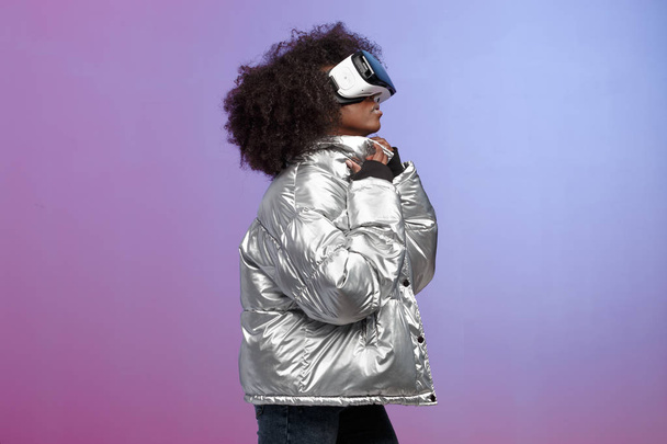 Trendy curly brown-haired girl dressed in a silver-colored jacket uses the virtual reality glasses in the studio on neon background - Foto, Bild