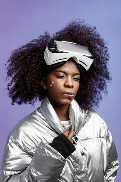 Modern curly brown-haired girl dressed in a silver-colored jacket  wearing on her head the virtual reality glasses poses in the studio on neon background - Foto, immagini