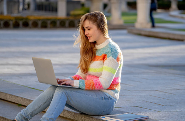 Pretty young cheerful teenager woman working or studying on laptop computer sitting happy and on stairs in city outdoors urban background. In student lifestyle Online education and Technology concept. - 写真・画像