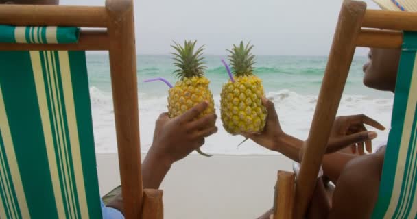 Rear view of African american couple toasting pineapple juice on the beach. They are relaxing on a sun loungers 4k  - Záběry, video