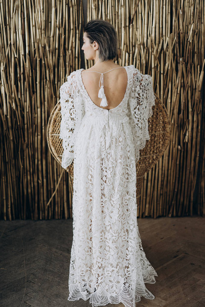 πίσω όψη της γυναίκας στο στούντιο φορώντας λευκό φόρεμα σε στυλ Μπόχο στέκεται ενάντια στο φόντο μπαμπού  - Φωτογραφία, εικόνα