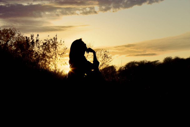 Silhouette einer jungen Frau, die bei Sonnenuntergang auf einem Hügel sitzt, ein Mädchen, das im Herbst auf dem Feld spaziert - Foto, Bild