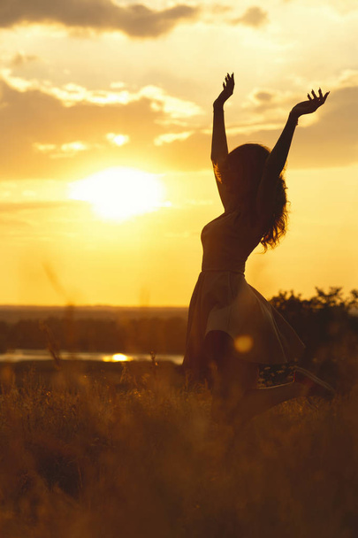Silhouette eines schönen Mädchens, das in einem Kleid bei Sonnenuntergang auf einem Feld tanzt und sich dreht, eine glückliche junge Frau, die die Natur genießt - Foto, Bild