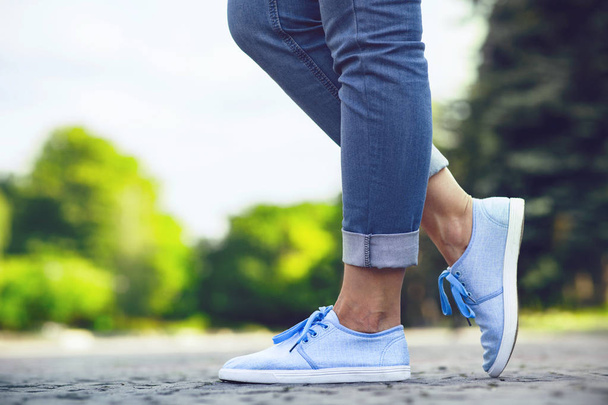 legs of a girl in jeans and blue sneakers on a sidewalk tile, a young woman strolling in a summer park - Fotó, kép