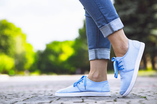 legs of a girl in jeans and blue sneakers on a sidewalk tile, a young woman strolling in a summer park - Foto, Imagen