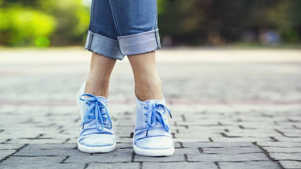 legs of a girl in jeans and blue sneakers on a sidewalk tile, a young woman strolling in a summer park - Fotoğraf, Görsel