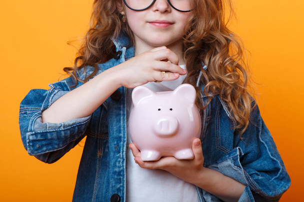 子供のポケットマネーの財政的概念。色付きのオレンジ色の背景にピギーバンクを持つ子供の女の子 - 写真・画像