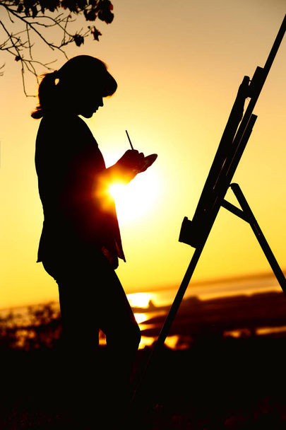 silueta mladé ženy, která kresla obrázek na stojanu na přírodě, Žena stojící pod stromem se štětcem a malířskou paletou, která se při západu slunce zabývá uměním - Fotografie, Obrázek