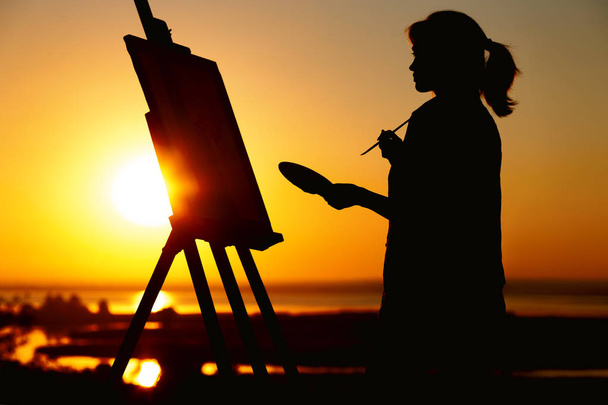 Silhouette einer Frau beim Bemalen eines Bildes mit Farben auf Leinwand auf einer Staffelei, Mädchen mit Pinsel und Palette in der Kunst auf einem Feld bei Sonnenuntergang - Foto, Bild