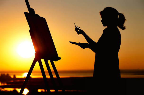 Silhouette einer Frau beim Bemalen eines Bildes mit Farben auf Leinwand auf einer Staffelei, Mädchen mit Pinsel und Palette in der Kunst auf einem Feld bei Sonnenuntergang - Foto, Bild