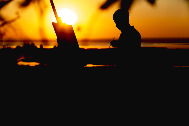 Silhouette eines Mannes beim Bemalen eines Bildes mit Farben auf Leinwand auf einer Staffelei über die Natur, Junge mit Pinsel und Palette bei Sonnenuntergang - Foto, Bild