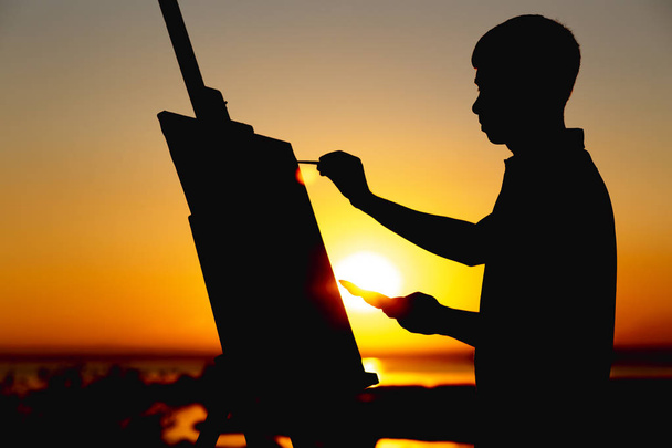 bir adamın silueti doğa üzerinde bir şelon üzerine tuval üzerine boyalar ile bir resim boyama, boya fırçası ve palet ile çocuk gün batımında sanat yapan - Fotoğraf, Görsel
