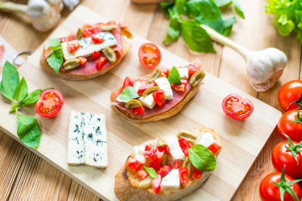 Bruschetta italiana tradicional com queijo azul, feta, tomates, folhas de manjericão, jamon em um fundo de madeira
. - Foto, Imagem