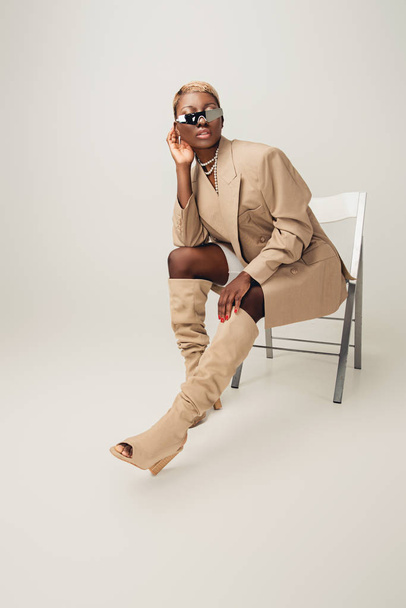 young african american woman in sunglasses and beige jacket sitting on chair on grey - 写真・画像