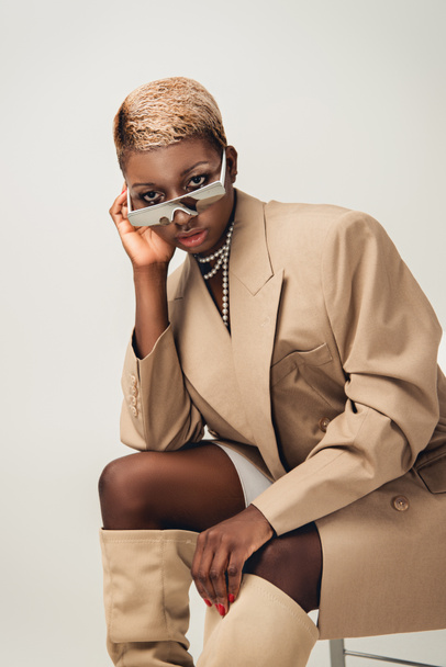 fashionable african american woman in sunglasses and beige jacket sitting on chair on grey - Fotó, kép