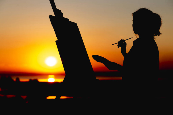 silueta ženy, která maluje obrázek na plátně na stojanu, dívka s kartáčkem na malování a paletou, která se zabývá uměním v terénu při západu slunce - Fotografie, Obrázek