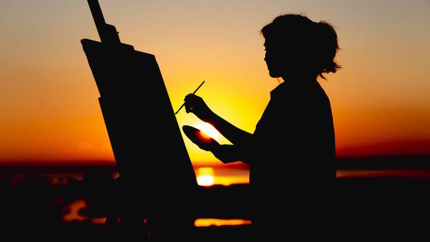 silhouette of a woman painting a picture on linen on an easel on the nature, painter face profile engaged in art in a field at sunset - Photo, Image