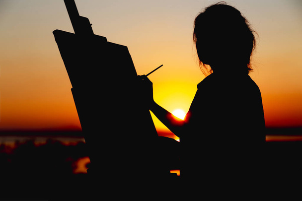 Silhouette einer Frau, die ein Bild auf Leinwand auf einer Staffelei malt, Mädchen mit Pinsel und Palette, die sich bei Sonnenuntergang auf einem Feld mit Kunst beschäftigt - Foto, Bild