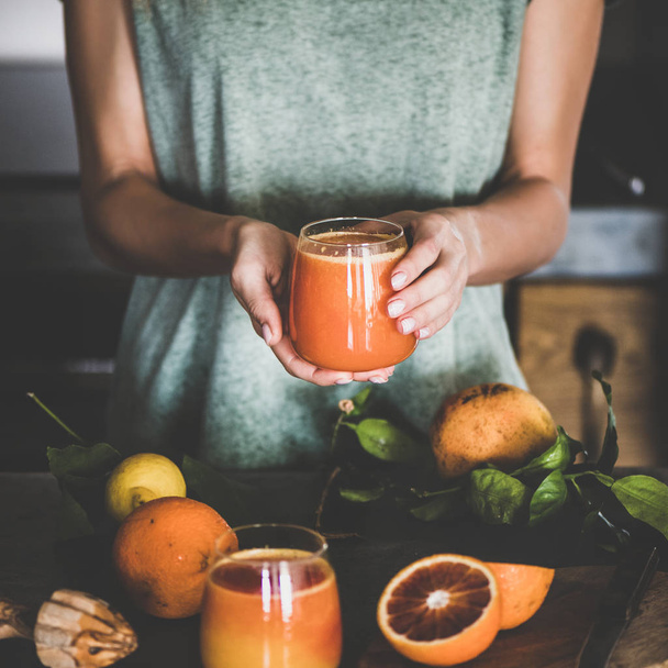 Young female holding glass of freshly squeezed blood orange juice or smoothie in hands near concrete kitchen counter, square crop. Healthy lifestyle, vegetarian, alkaline diet, spring detox concept - Photo, image