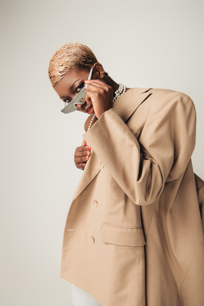 stylish african american girl with short hair posing in sunglasses and beige jacket isolated on grey - Foto, imagen