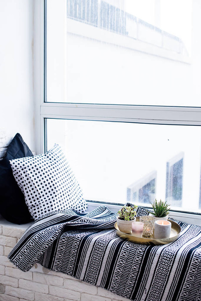 Apartamento real decoración interior, velas aromáticas y plantas en bandeja vintage con almohadas y manta en alféizar de ventana blanca
 - Foto, Imagen