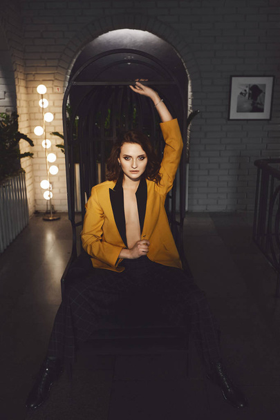 Sexy young woman in the yellow blazer on the naked body and in trendy checkered pants sits on a metal chair and posing at the dark interior - Φωτογραφία, εικόνα