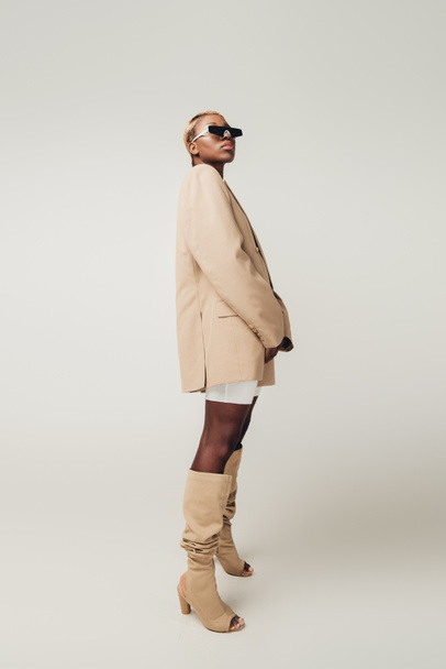 modne African American Girl stwarzających w okulary przeciwsłoneczne i beżowy kurtka izolowane na szarym - Zdjęcie, obraz