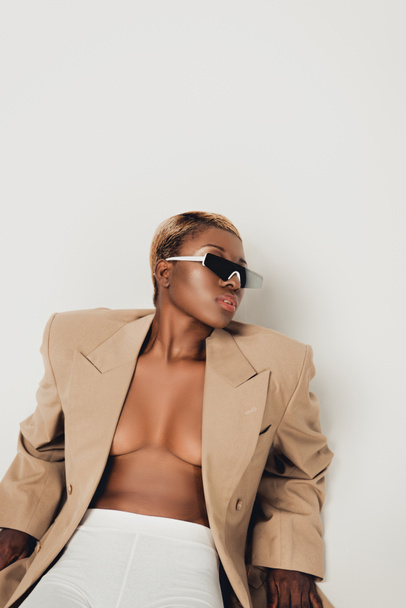 chica afroamericana elegante posando en gafas de sol y chaqueta beige aislado en gris
 - Foto, imagen