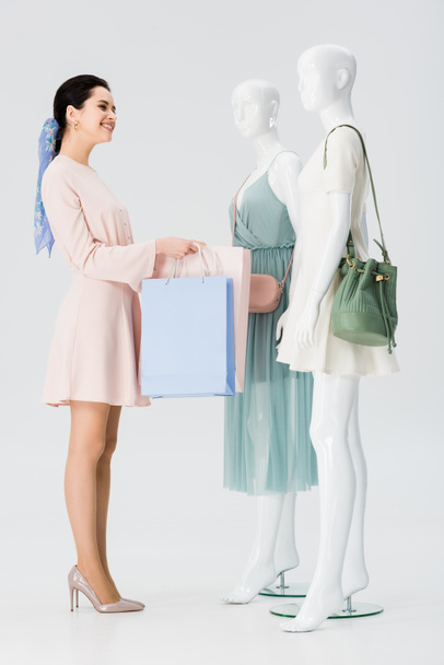 jovem sorridente com sacos de compras perto de manequins isolados em cinza
 - Foto, Imagem