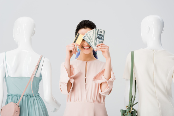 beautiful girl near mannequins covering face with money and credit card isolated on grey - Photo, Image