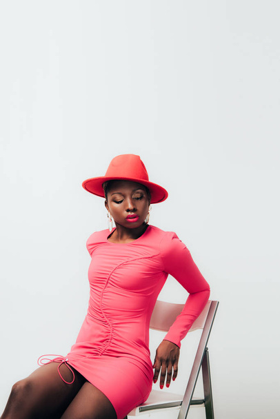 joven mujer afroamericana sensual en vestido rosa y sombrero posando en silla aislada en blanco
 - Foto, imagen