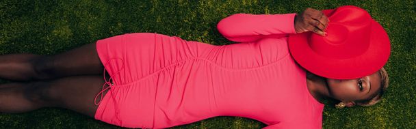 top view of elegant african american woman in pink dress lying with hat on green grass - Photo, Image