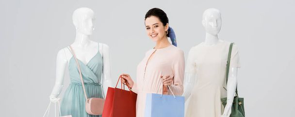 panoramic shot of beautiful young woman with shopping bags near mannequins isolated on grey - Foto, imagen