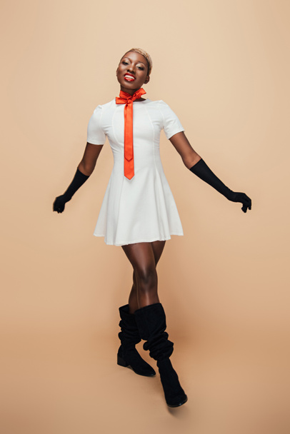 χαμογελαστή Αφρικανική Αμερικάνικη κοπέλα που ποζάρει σε λευκό φόρεμα, κόκκινο κασκόλ και μαύρα γάντια στο μπεζ - Φωτογραφία, εικόνα