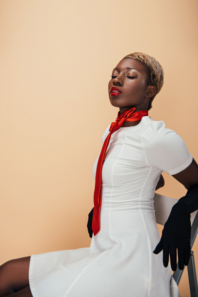 白いドレス、赤いスカーフ、ベージュに隔離された黒い手袋でポーズをとるアフリカ系アメリカ人モデル - 写真・画像